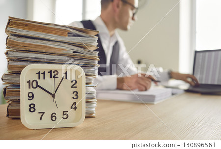 Clock and stack of paperwork on office table, with man working on laptop in background 130896561