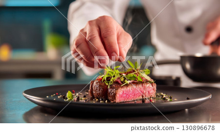 The chef decorates a medium-rare beef steak with fresh microgreens on a black plate, exquisite cuisine for gourmets 130896678