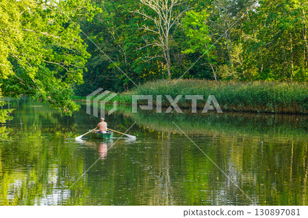 Man rowing boat on calm forest river with green reflections of trees in peaceful summer landscape 130897081