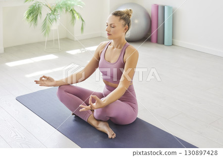 Sporty yogi woman sitting cross-legged on yoga mat in lotus pose, meditating with her eyes closed Sporty yogi woman sitting cross-legged on yoga mat in lotus pose, meditating with her eyes closed 130897428