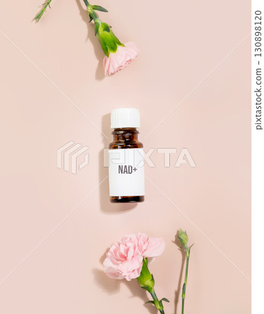 NAD essence in vial with white cap surrounded by pink carnations on nude pastel background. Concept of NAD beauty products, cellular beauty, anti-aging trends, and modern wellness products NAD essence in vial with white cap surrounded by pink carnations on nude pastel background. Concept of NAD beauty products, cellular beauty, anti-aging trends, and modern wellness products 130898120