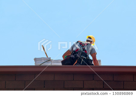 Professional Roofer Installing Asphalt Shingles on Residential Roof with Safety Equipment 130898869