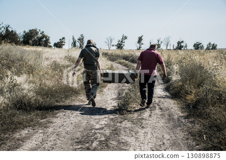 Back view of two men walking on rural dirt road carrying heavy metal box, concept of war or military mission Back view of two men walking on rural dirt road carrying heavy metal box, concept of war or military mission 130898875