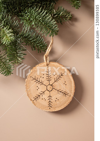Christmas toy made of decorative wooden slice with ornament snowflake hangs evergreen branches on beige background. Xmas vertical card. View from above. Christmas toy made of decorative wooden slice with ornament snowflake hangs evergreen branches on beige background. Xmas vertical card. View from above. 130899385