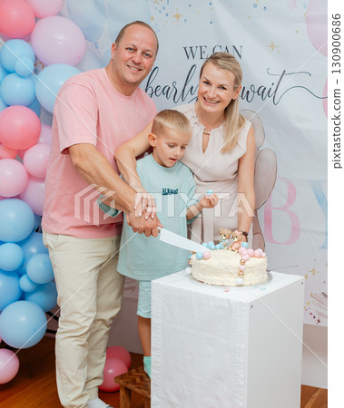 Parents and child holding knife to cut cake at family party at gender reveal celebration. Pink and blue balloons in background, festive lifestyle editorial concept 130900686