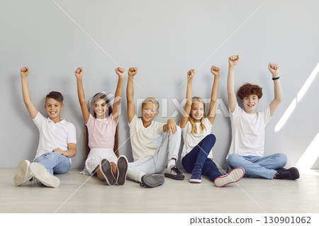 Group of school children sitting in a row on the floor on gray background with hands up in fist. Group of school children sitting in a row on the floor on gray background with hands up in fist. 130901062