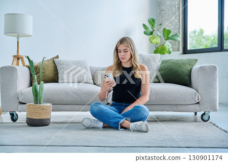 Serious young woman with smartphone sitting at home near sofa Serious young woman with smartphone sitting at home near sofa 130901174
