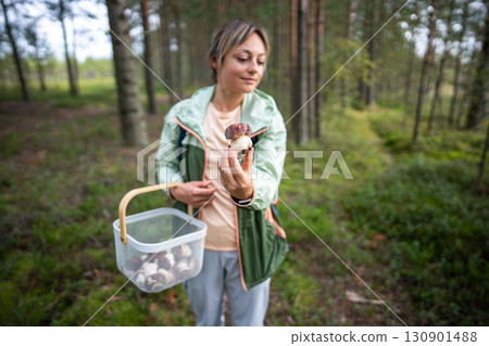Satisfied woman gathering mushrooms in autumn forest with basket, holding fresh bolete in hand Satisfied woman gathering mushrooms in autumn forest with basket, holding fresh bolete in hand 130901488