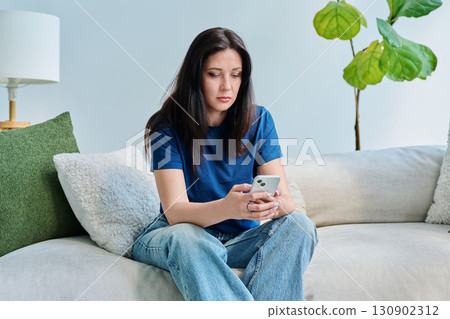 Serious young woman sitting at home on the couch using smartphone Serious young woman sitting at home on the couch using smartphone 130902312