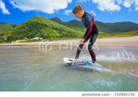 Young boy skimboarding in shallow water on a sunny beach day. Wearing wetsuit, balancing on board with determination and focus. Perfect image for themes of childhood adventure, active lifestyle, Young boy skimboarding in shallow water on a sunny beach day. Wearing wetsuit, balancing on board with determination and focus. Perfect image for themes of childhood adventure, active lifestyle, 130902330