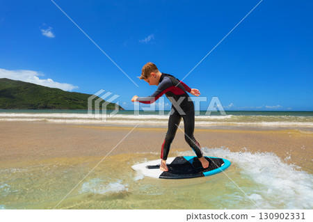 Young boy skimboarding in shallow water on a sunny beach day. Wearing wetsuit, balancing on board with determination and focus. Perfect image for themes of childhood adventure, active lifestyle, 130902331