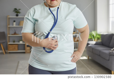 Fat woman standing at home and listening heartbeat on stethoscope after doing exercise. Fat woman standing at home and listening heartbeat on stethoscope after doing exercise. 130902380