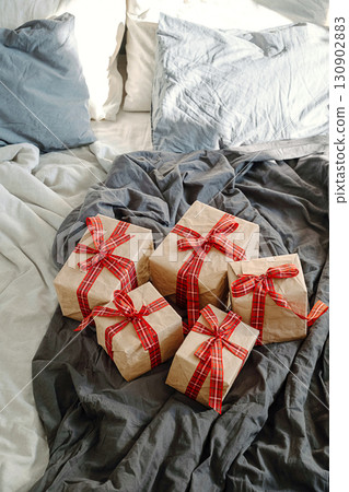 Wrapped brown paper gift boxes with red plaid ribbon on grey rumpled bed. Christmas morning present surprise. Wrapped brown paper gift boxes with red plaid ribbon on grey rumpled bed. Christmas morning present surprise. 130902883