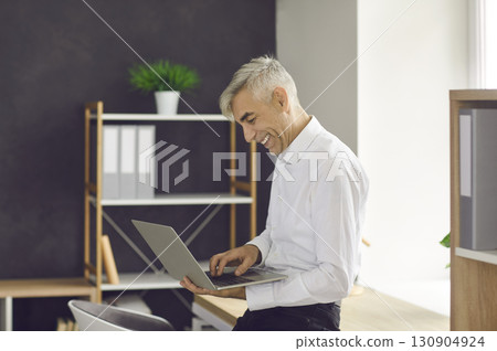 Happy businessman standing by office desk, holding laptop and doing some work online 130904924