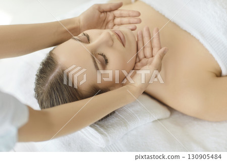 Beautician hands massaging woman face, female client lying relaxing with eyes closed in spa saloon Beautician hands massaging woman face, female client lying relaxing with eyes closed in spa saloon 130905484