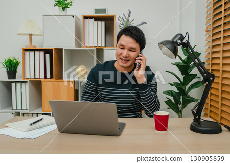 happy man talking with smartphone while working with laptop computer at home office happy man talking with smartphone while working with laptop computer at home office 130905859