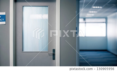 A closed, gray hospital or office door with a vertical glass window is seen in a clean, empty hallway. 130905956