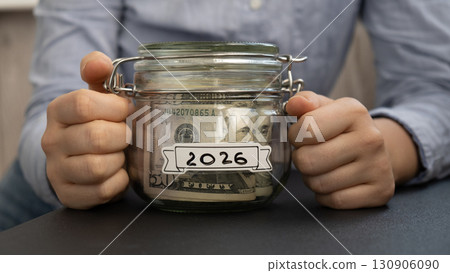 Unrecognizable woman holding Saving Money In Glass Jar filled with Dollars banknotes. 2026 year transcription in front of jar. Managing personal finances extra income for future insecurity Unrecognizable woman holding Saving Money In Glass Jar filled with Dollars banknotes. 2026 year transcription in front of jar. Managing personal finances extra income for future insecurity 130906090