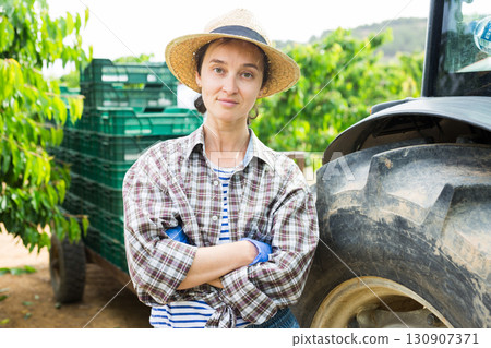 Portrait of female farmer stands next to tractor in an orchard Portrait of female farmer stands next to tractor in an orchard 130907371