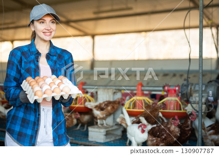 Smiling european woman in plaid shirt and cap collecting eggs in chicken farm 130907787