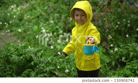 child in yellow raincoat with hood stands in the rain filling a small watering can with rainwater in front of a green garden 130908741