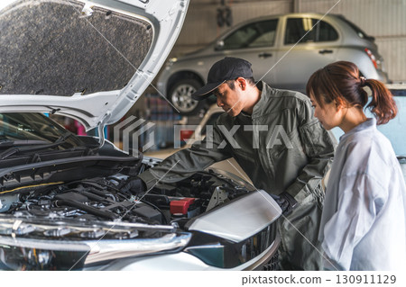 A mechanic explaining to a female customer in the car's engine room, a male mechanic staff member A mechanic explaining to a female customer in the car's engine room, a male mechanic staff member 130911129