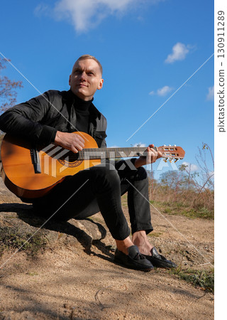 Beautiful blonde man playing acoustic guitar and singing relaxing enjoying outdoors on sunny autumn day. Artist learning and practicing playing musical instrument. Fall leaves autumnal 130911289