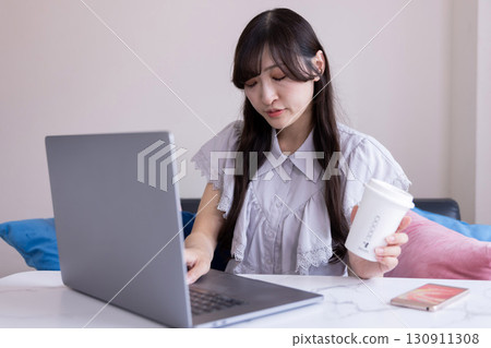 A Japanese woman with a serious expression working on a PC in the living room with a mug in her hand 130911308