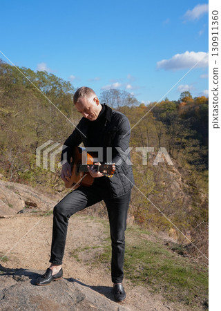 Portrait of smiling caucasian artist plays wooden acoustic guitar in autumnal park. Young songwriter plays string musical instrument hobby outside in nature fall time. Audio music healing 130911360