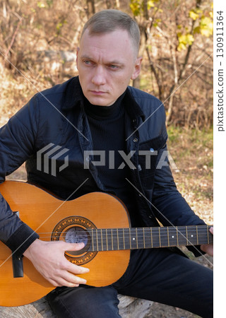 Portrait of smiling caucasian artist plays wooden acoustic guitar in autumnal park. Young songwriter plays string musical instrument hobby outside in nature fall time. Audio music healing 130911364