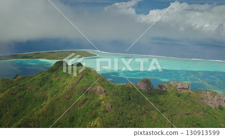 Aerial drone capturing a breathtaking tropical island with lush vegetation green hill mountains, surrounded by a vibrant turquoise lagoon and protected by a coral reef in French Polynesia 130913599