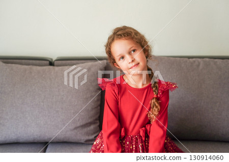 A young girl in a red festive dress sits on a gray couch, looking calmly at the camera 130914060