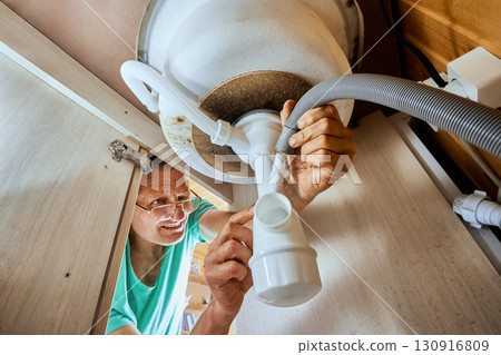 Maintenance worker installs bottle trap inlet pipe under kitchen sink securing connection with overflow hose and dishwasher waste hose during assembly. 130916809