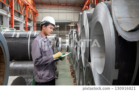 A worker dressed in hardhat and  protective glove, stand in a factory check material while holding document clipboard. The environment is organized with rolls of metal. 130916821