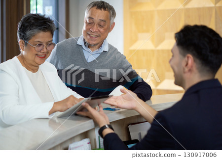 Asian senior couple is engaging with hotel staff at the reception desk, receiving assistance as they check in. The setting is modern and welcoming, creating a friendly atmosphere. Asian senior couple is engaging with hotel staff at the reception desk, receiving assistance as they check in. The setting is modern and welcoming, creating a friendly atmosphere. 130917006