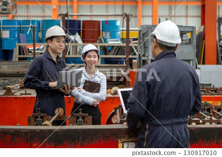 Three workers in a factory discuss a project using laptop computer. Everyone wearing protective hardhat. Atmosphere in manufactory with large machinery. 130917010