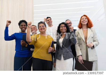 A diverse group of professional stand together in a modern office, celebrating with raised fist and smile. The atmosphere is lively and filled with positivity, showcasing teamwork and achievement. 130917397