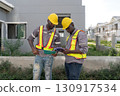 Two construction worker in a helmet and safety vest review plan on tablet at a building site. They focus intently on their task while discussing project detail. 130917534