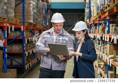 Business woman in suit and safety helmet review inventory record on laptop computer. Stand in a well-organized warehouse. Shelve are lined with various product, indicating a busy working environment Business woman in suit and safety helmet review inventory record on laptop computer. Stand in a well-organized warehouse. Shelve are lined with various product, indicating a busy working environment 130917539