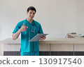 A young male healthcare worker in teal scrub stand at a reception desk in a well-lit clinic. He is holding a clipboard and looking confidently at the camera, ready for consultation. 130917540