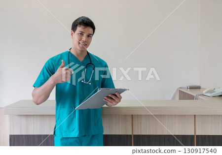 A young male healthcare worker in teal scrub stand at a reception desk in a well-lit clinic. He is holding a clipboard and looking confidently at the camera, ready for consultation. A young male healthcare worker in teal scrub stand at a reception desk in a well-lit clinic. He is holding a clipboard and looking confidently at the camera, ready for consultation. 130917540
