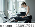 A woman take a break while exercising on a treadmill, catching her breath in a bright, contemporary gym setting with large window. 130917541