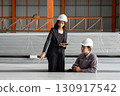 Two worker in hardhat talk in a warehouse. They discuss plans while standing near large, wrapped material. 130917542