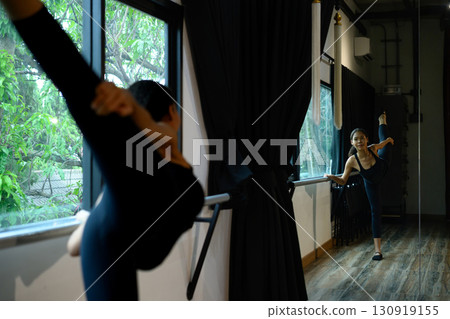 Young ballerina practicing leg stretch at barre in dance studio with mirror reflection Young ballerina practicing leg stretch at barre in dance studio with mirror reflection 130919155