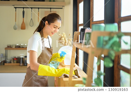 Young woman smiling while cleaning kitchen shelves with spray bottle and cloth Young woman smiling while cleaning kitchen shelves with spray bottle and cloth 130919163