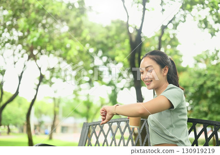 Smiling woman sitting on a park bench looking at her wristwatch, enjoying a relaxed moment in nature Smiling woman sitting on a park bench looking at her wristwatch, enjoying a relaxed moment in nature 130919219