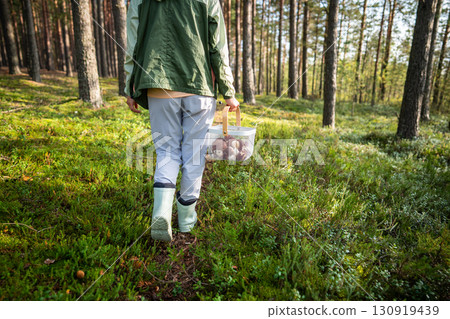 Woman walking through forest on trail carrying basket of mushrooms, fall nature hobby, harvest time 130919439