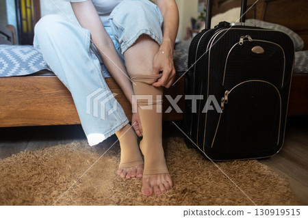 Woman putting on compression stockings before a long trip, suitcase in the background. Concept of leg protection during travel, varicose veins and deep vein thrombosis on long flights or car journeys Woman putting on compression stockings before a long trip, suitcase in the background. Concept of leg protection during travel, varicose veins and deep vein thrombosis on long flights or car journeys 130919515