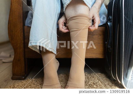 Woman putting on compression stockings before a long trip, suitcase in the background. Concept of leg protection during travel, varicose veins and deep vein thrombosis on long flights or car journeys Woman putting on compression stockings before a long trip, suitcase in the background. Concept of leg protection during travel, varicose veins and deep vein thrombosis on long flights or car journeys 130919516