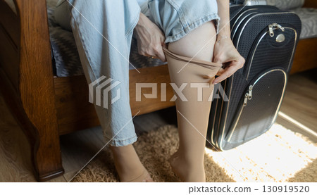 Woman putting on compression stockings before a long trip, suitcase in the background. Concept of leg protection during travel, varicose veins and deep vein thrombosis on long flights or car journeys Woman putting on compression stockings before a long trip, suitcase in the background. Concept of leg protection during travel, varicose veins and deep vein thrombosis on long flights or car journeys 130919520